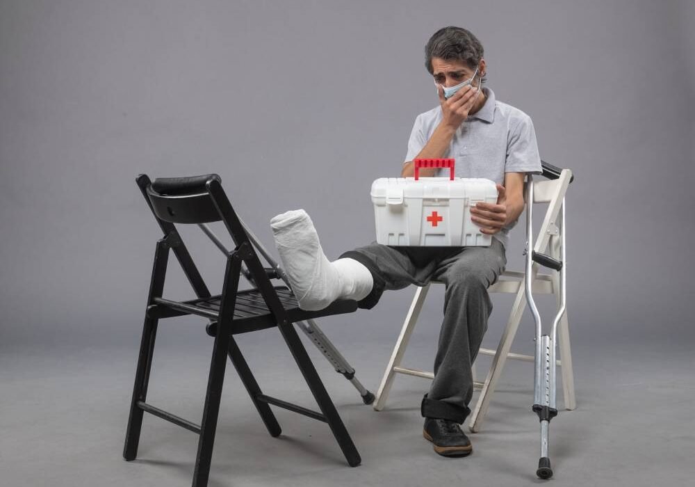 front view young male with mask and sitting with bandaged broken foot on grey desk twist male broke pain foot leg